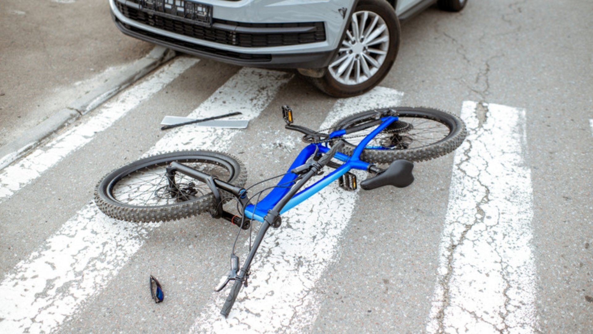 A blue bicycle lies on a crosswalk in front of a stopped silver car