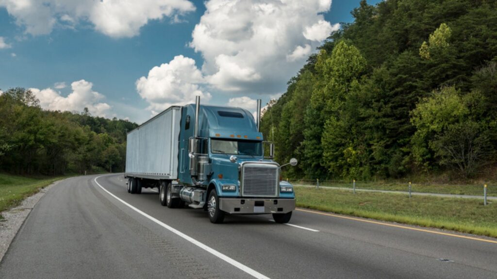 A blue semi truck pulling a white trailer along a two-lane highway past wooded hills